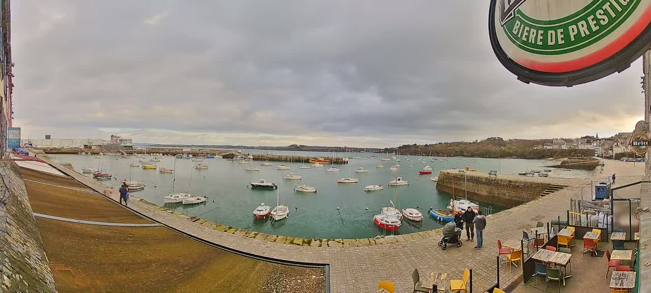 A harbor filled with numerous sailboats and motorboats under an overcast sky, viewed from a paved area with outdoor seating near a building.