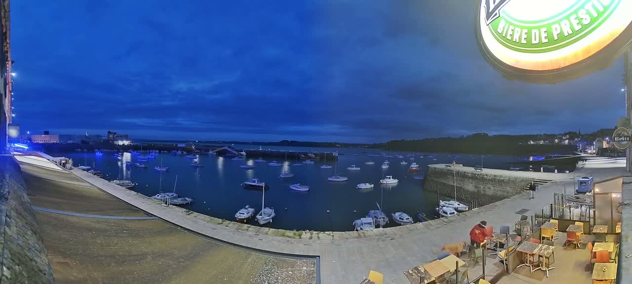Under a twilight sky, numerous small boats are moored in a harbor next to an outdoor seating area of a restaurant.