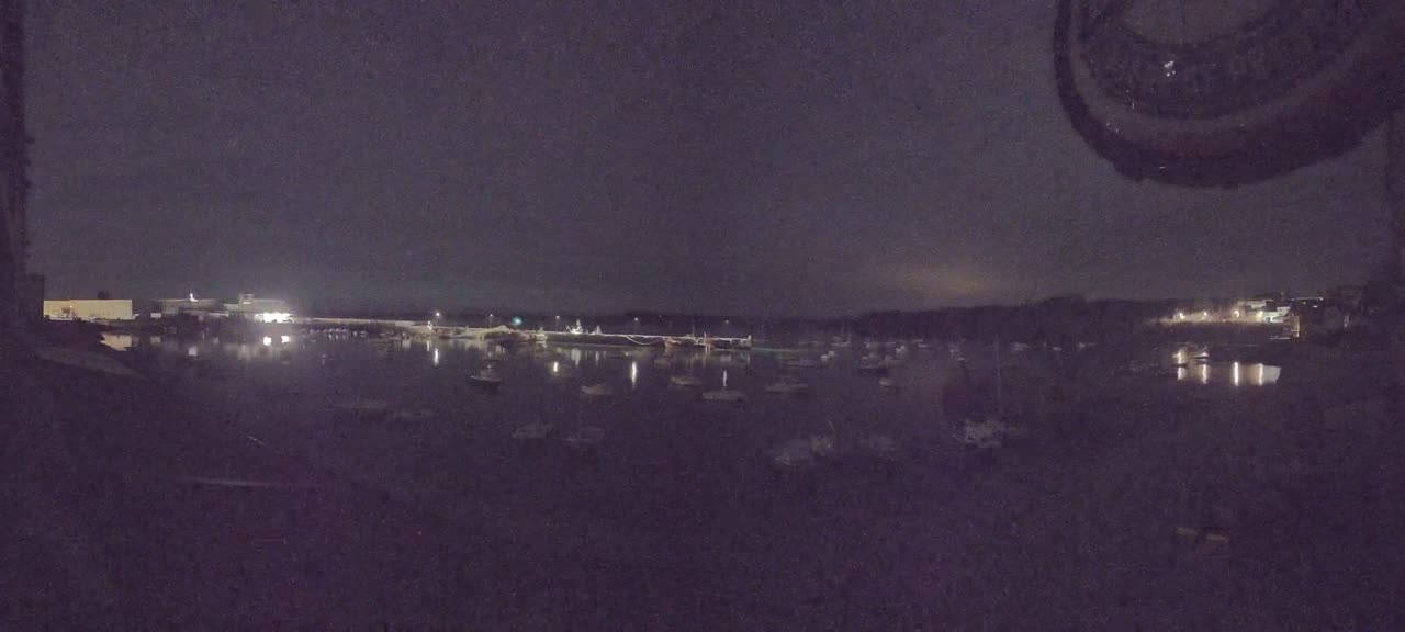 At night, numerous boats are moored in a calm body of water, illuminated by faint lights from a nearby shoreline.