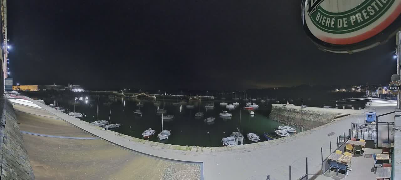 A nighttime view of a harbor filled with small boats, illuminated by artificial lights under a dark sky.