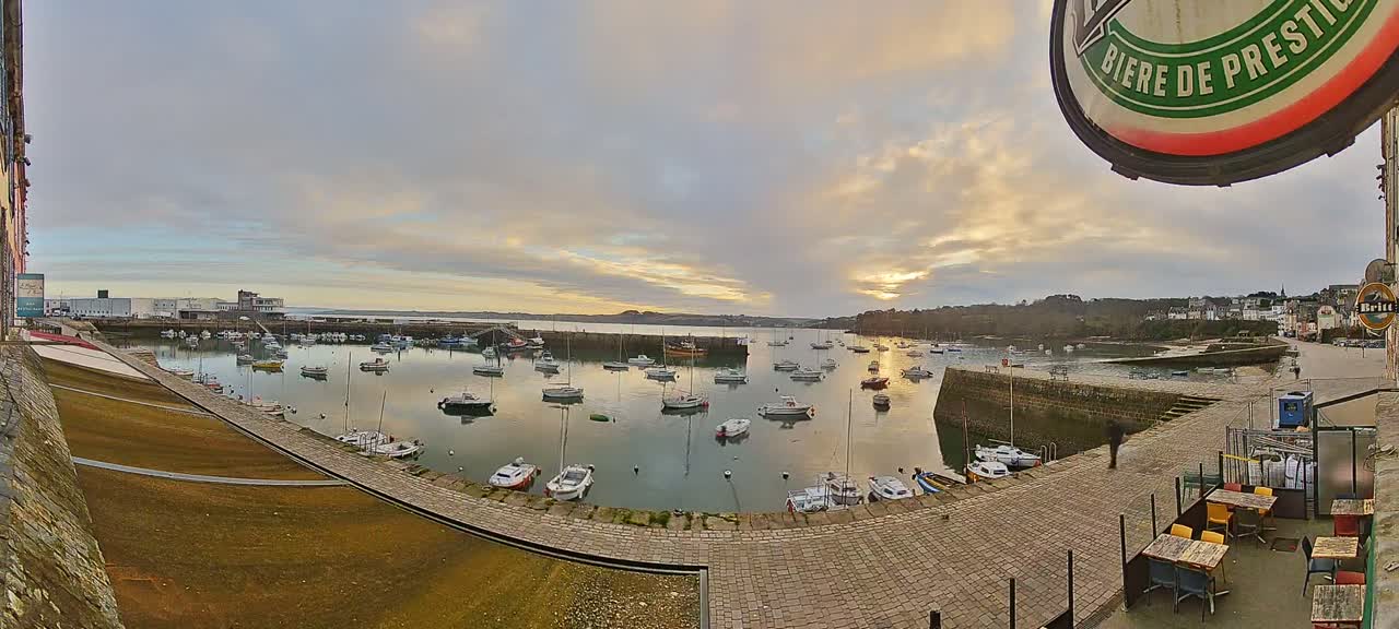 A harbor filled with numerous boats is seen at sunset under a cloudy sky, with a stone walkway and outdoor seating area in the foreground.