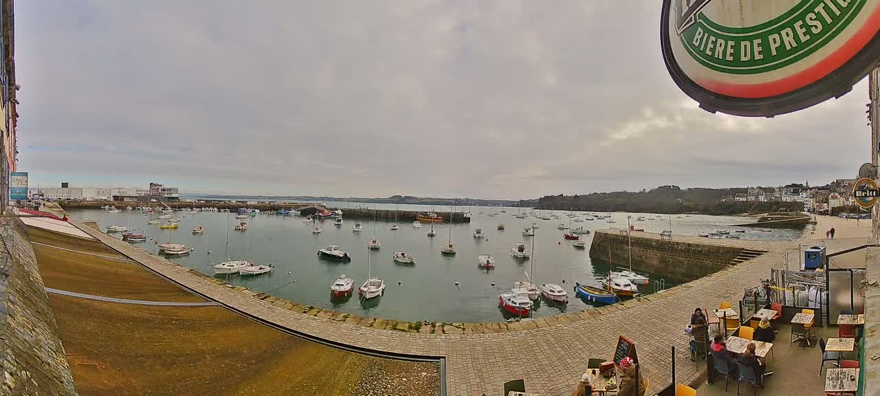 A harbor filled with numerous small boats is seen under an overcast sky, with a stone walkway and outdoor seating area in the foreground.
