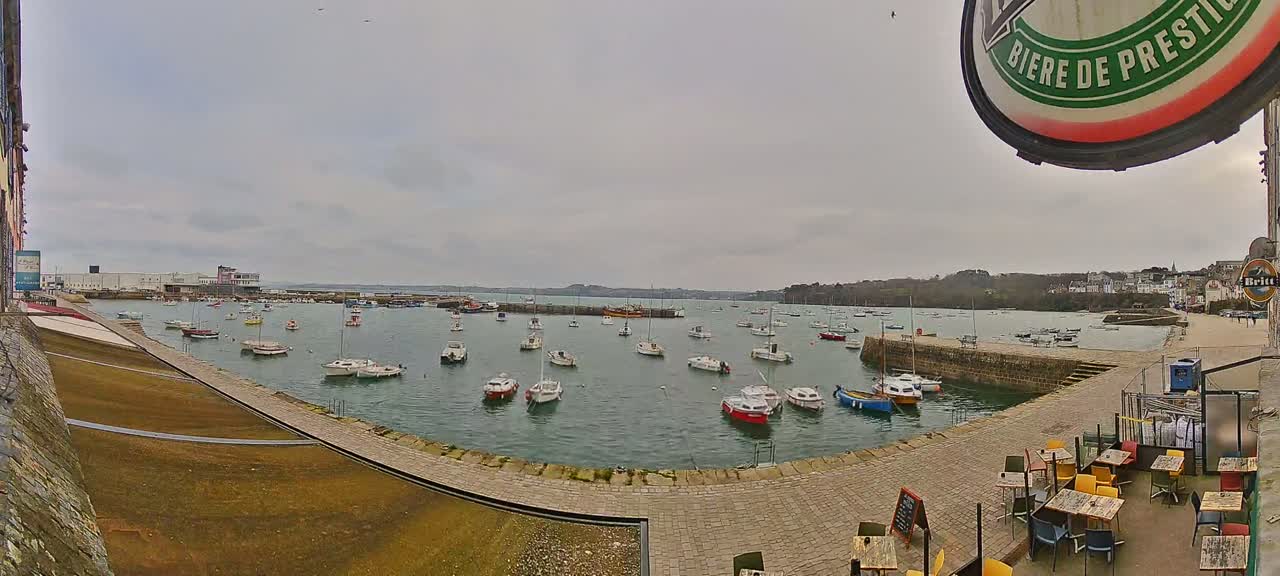 Numerous small boats are moored in a calm harbor on an overcast day, with a paved waterfront area and outdoor seating visible in the foreground.