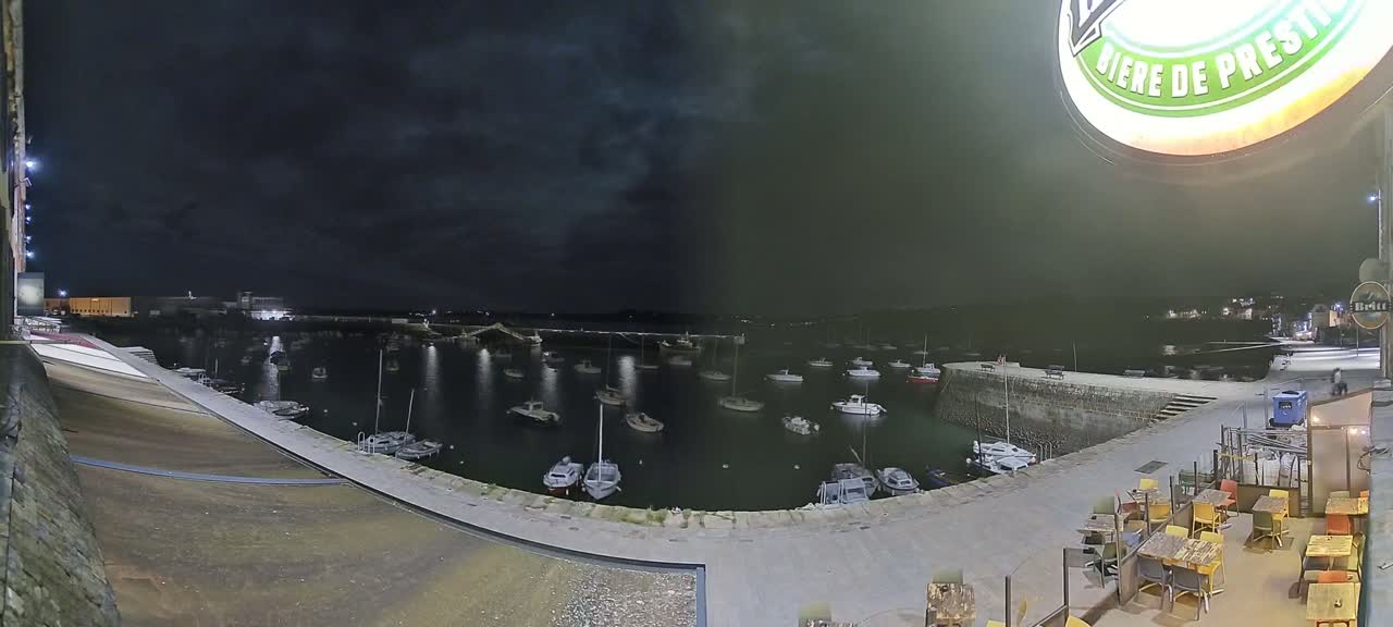 A nighttime view of a harbor filled with boats, seen from an outdoor patio area under a dark, cloudy sky.