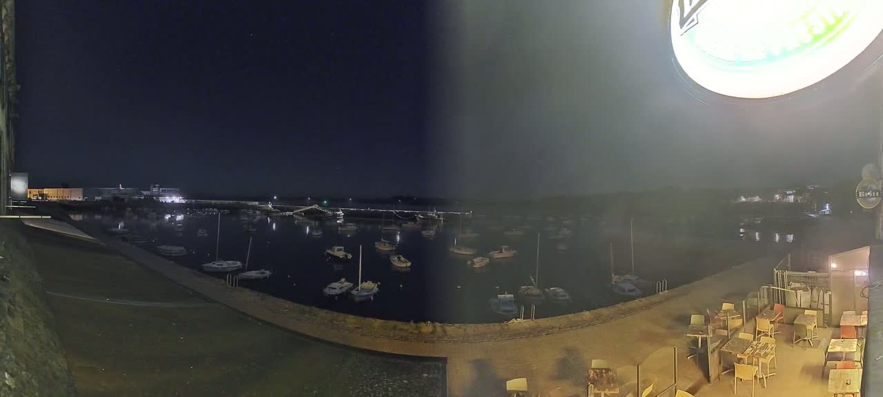 At night, a calm body of water with many boats is seen from an elevated viewpoint overlooking a patio area with outdoor seating.