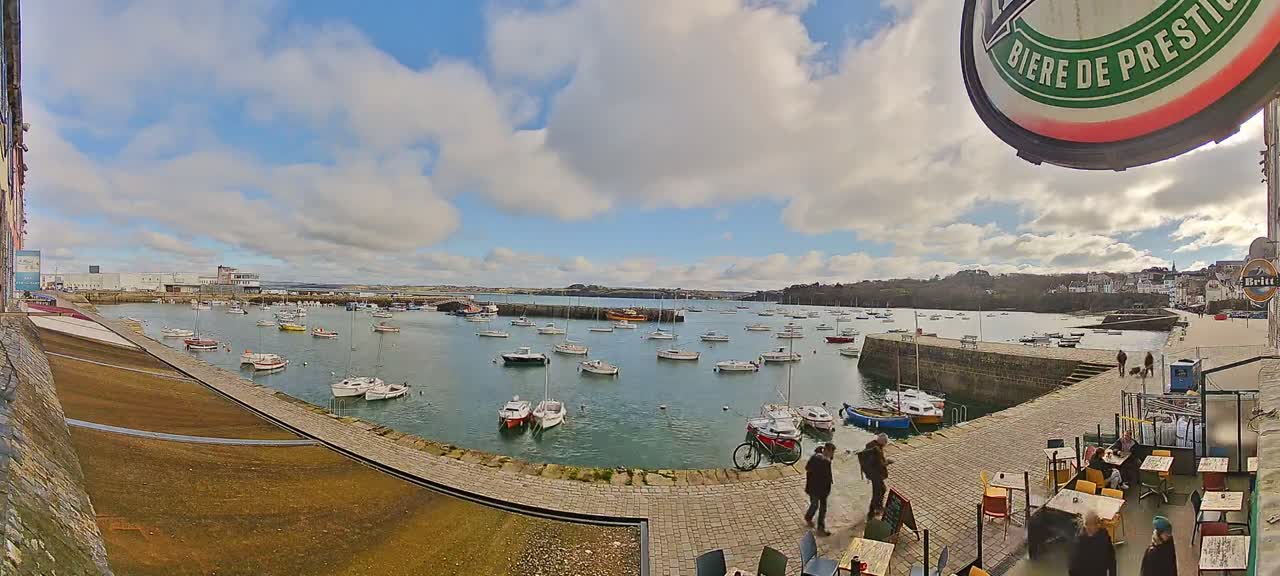 A harbor filled with numerous small boats is viewed from a paved area with outdoor seating under a partly cloudy sky.