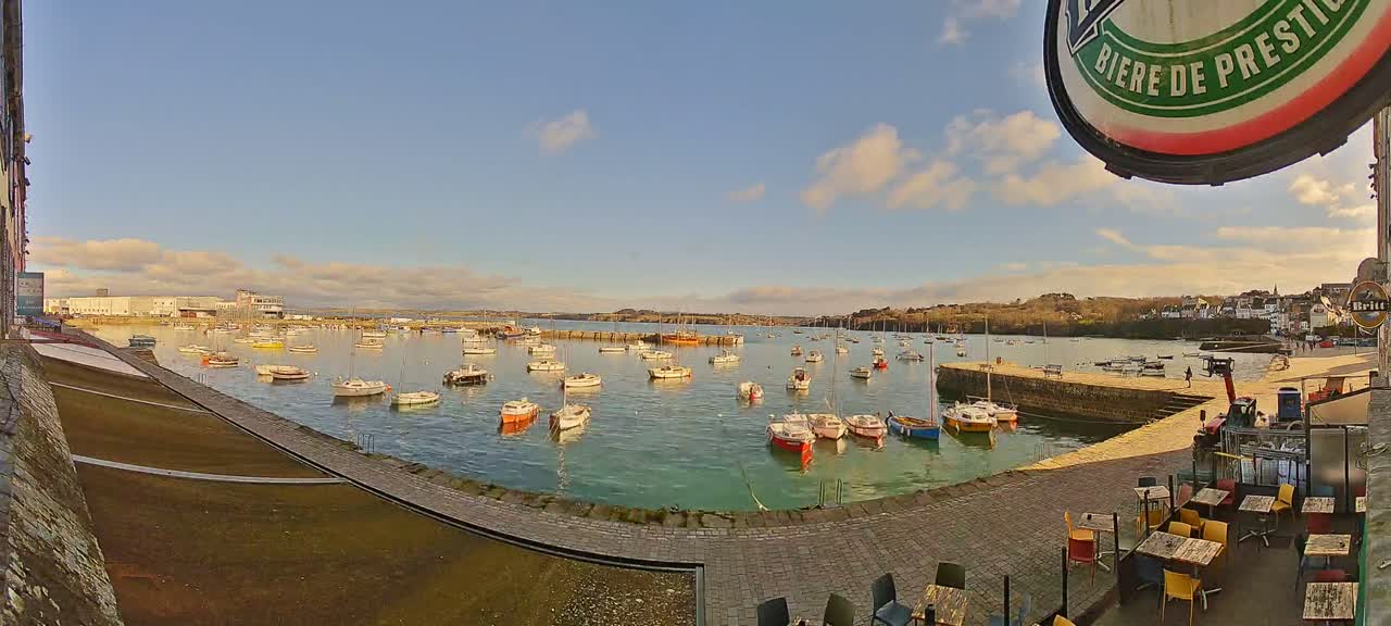 A harbor filled with numerous small boats on a partly sunny day, with a paved waterfront area and outdoor seating visible in the foreground.
