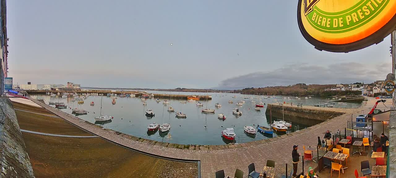 A harbor filled with numerous small boats under a mostly cloudy sky, with a stone walkway leading to an outdoor seating area of a restaurant.