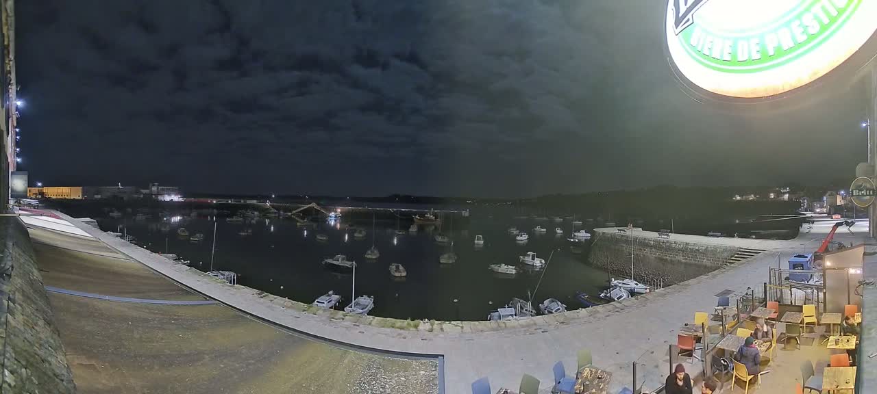 A nighttime view of a harbor filled with small boats under a cloudy sky, with an outdoor seating area visible in the foreground.