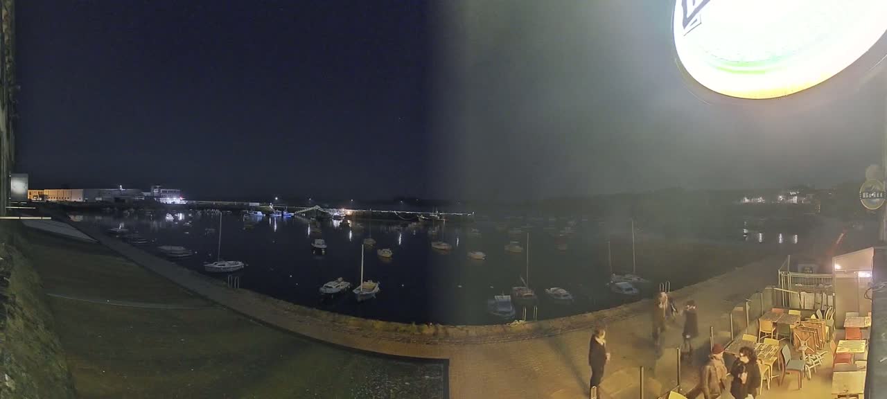 A nighttime view of a harbor filled with boats, seen from a slightly elevated vantage point overlooking a waterfront walkway with several people walking and an outdoor patio area with tables and chairs.