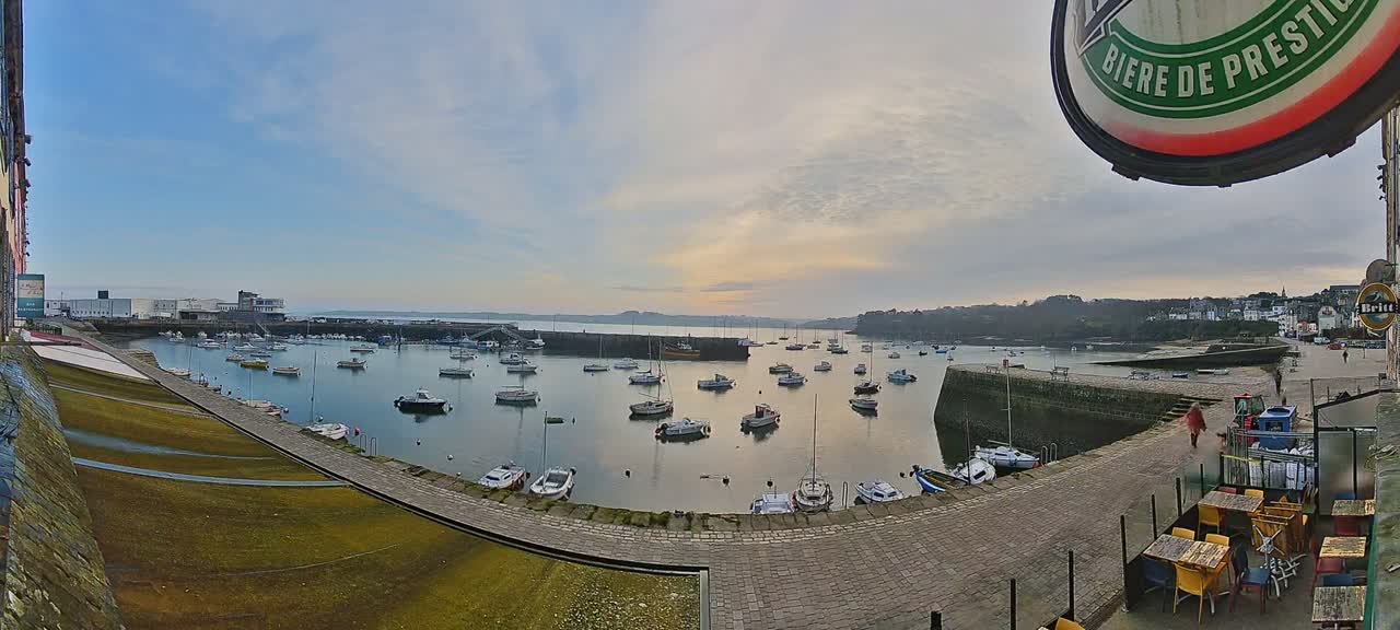 A harbor filled with numerous boats is seen under a partly cloudy sky at sunset, with a paved walkway and outdoor seating area in the foreground.
