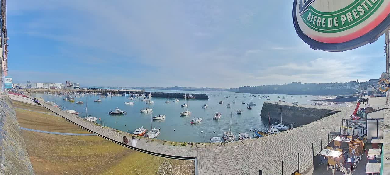 A sunny day reveals a harbor filled with numerous sailboats and motorboats, nestled within a stone-walled cove next to a paved walkway and outdoor seating area.