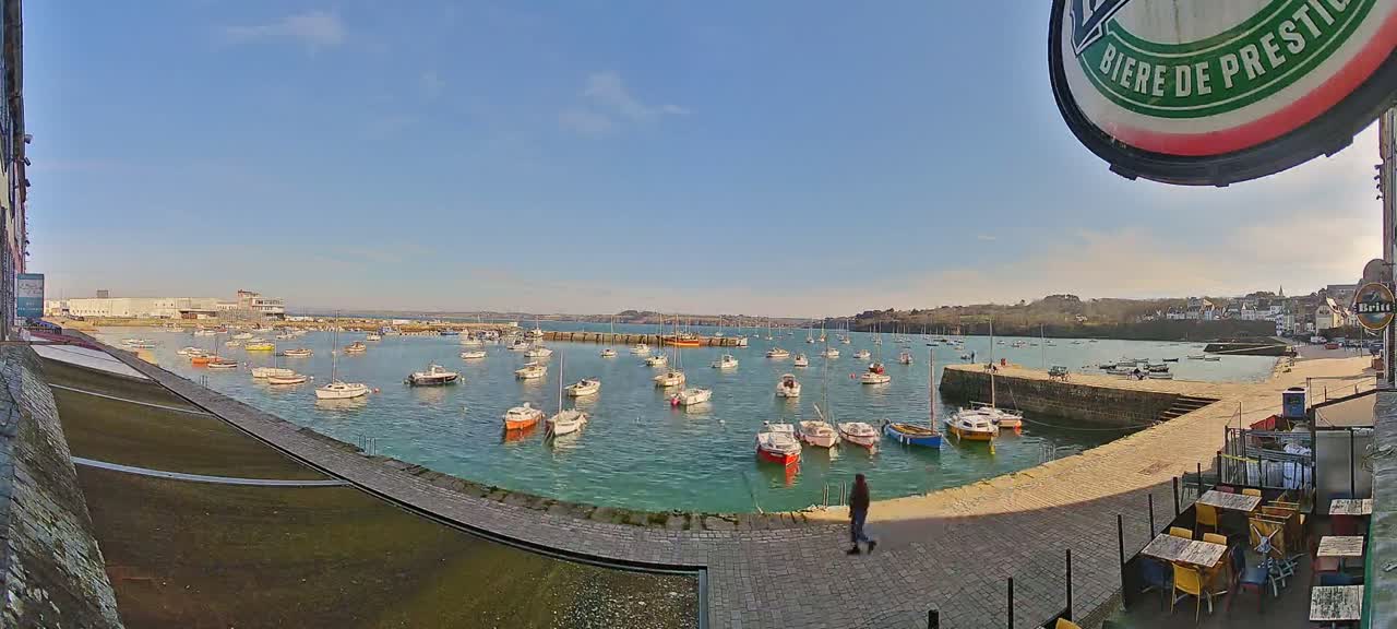 A sunny day reveals a harbor filled with numerous small boats, a stone walkway leading to outdoor seating, and a person walking near the water.