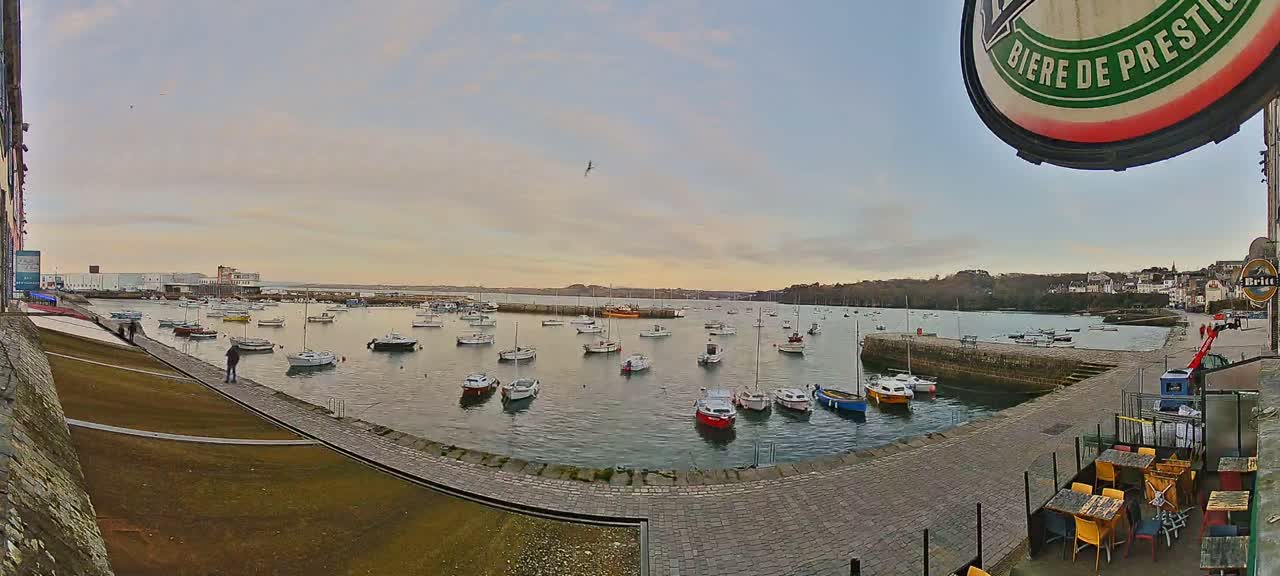 Many small boats are moored in a calm harbor under a partly cloudy, pale sunset sky.
