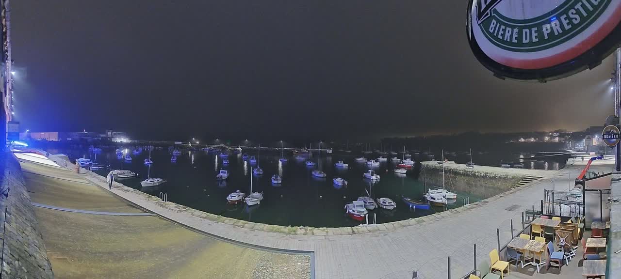 At night, a harbor filled with numerous boats is seen from a waterfront patio with outdoor seating.