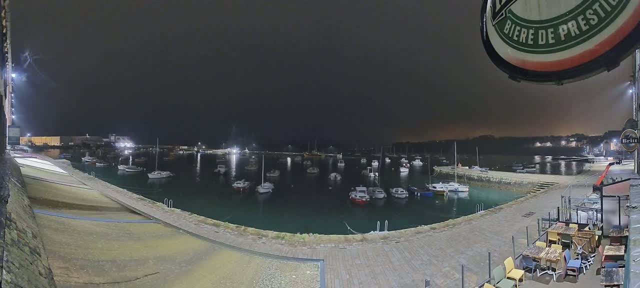 A nighttime view of a harbor filled with boats, seen from a patio with outdoor seating.
