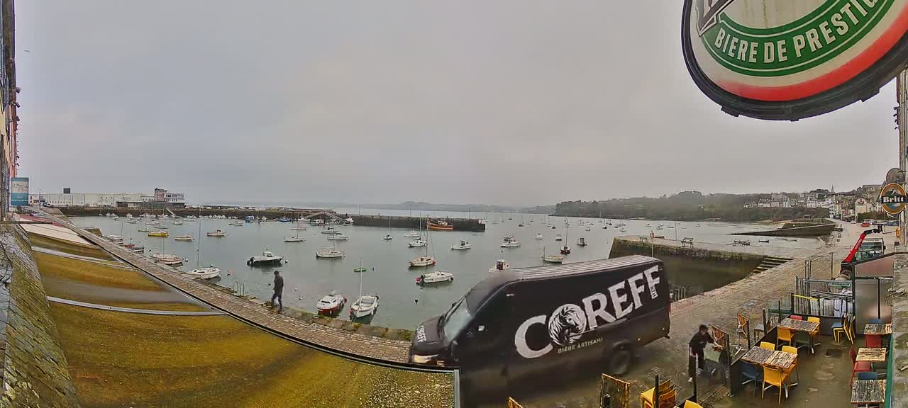 A harbor filled with numerous boats is viewed from a waterfront patio on an overcast day, with a van parked nearby.