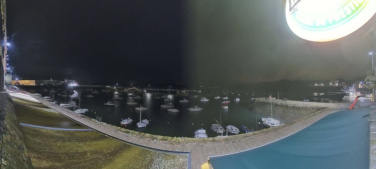 A nighttime harbor scene shows numerous boats moored in calm water under a dark sky.