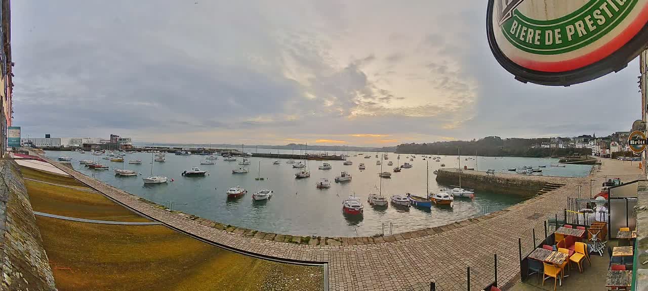 A harbor filled with numerous small boats is seen under an overcast sky at sunset, with a stone walkway and outdoor seating area in the foreground.