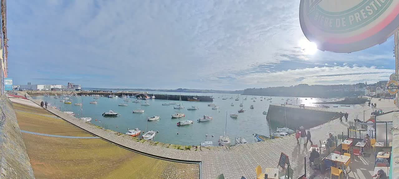 A sunny day overlooking a harbor filled with numerous boats, a paved walkway with people walking and outdoor seating areas.