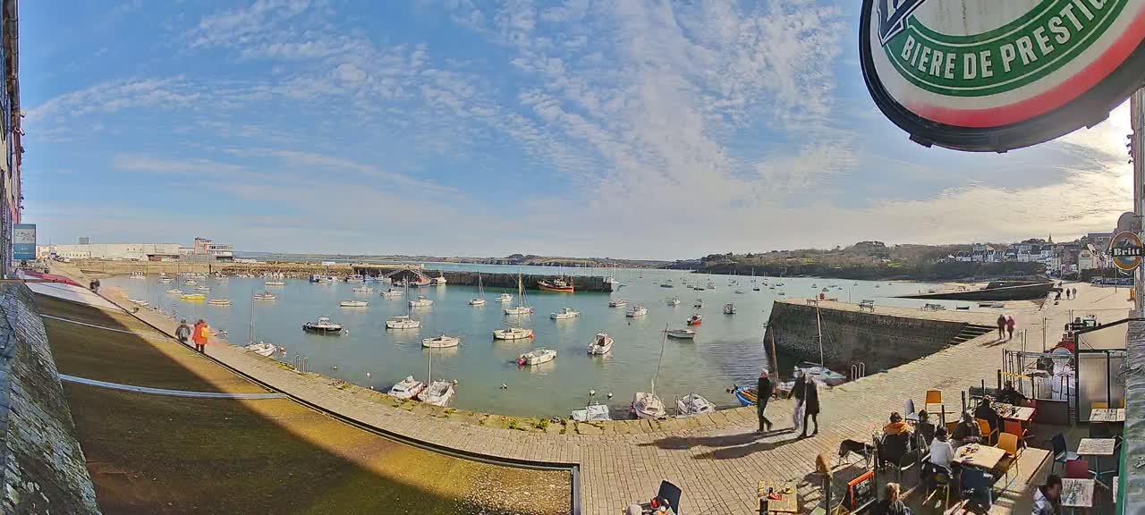 A sunny harbor scene with numerous boats at anchor, people walking along a stone walkway, and an outdoor cafe with patrons.