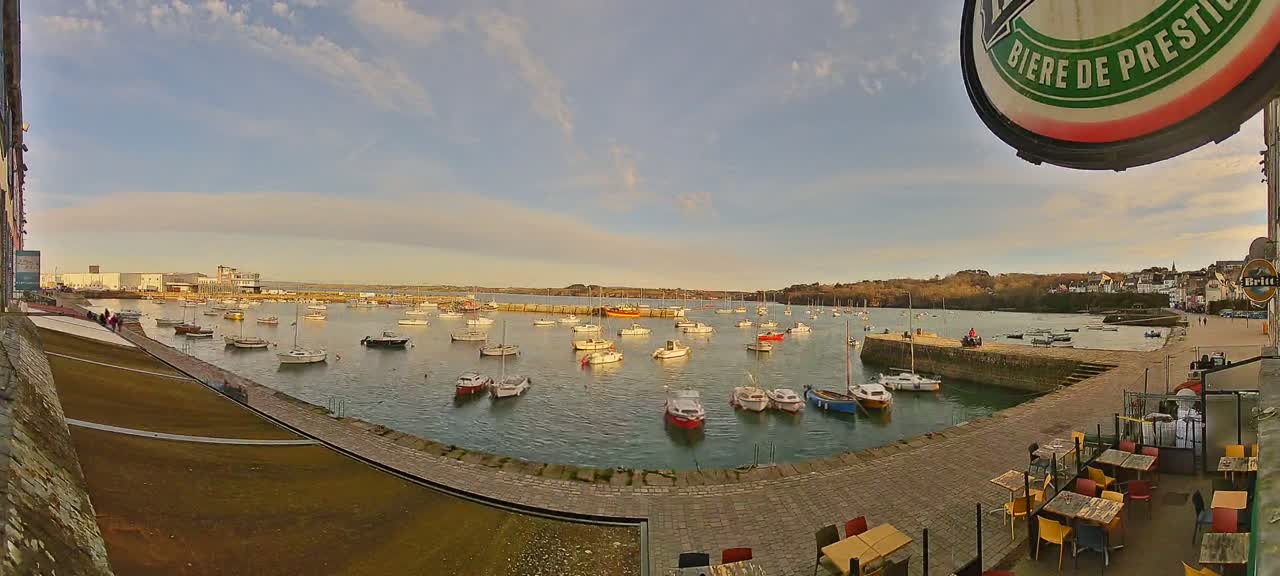 A harbor filled with numerous small boats under a partly cloudy sky, viewed from a paved area with outdoor seating.