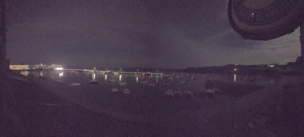 Many boats are moored in a dark, calm harbor at night, under a mostly cloudy sky.