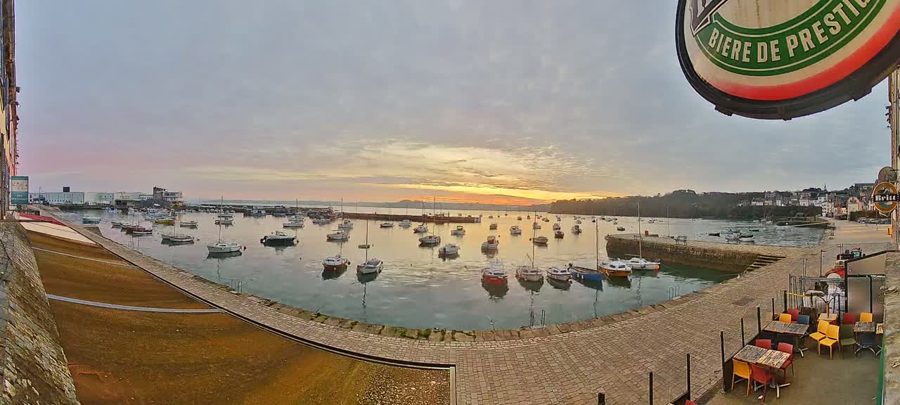Many small boats are moored in a harbor at sunrise under a mostly cloudy sky, with a paved area and outdoor seating visible in the foreground.