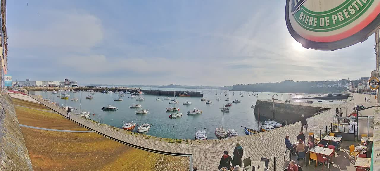 A sunny harbor scene shows numerous small boats in calm water, a paved waterfront area with people sitting at outdoor tables, and buildings along the shoreline.