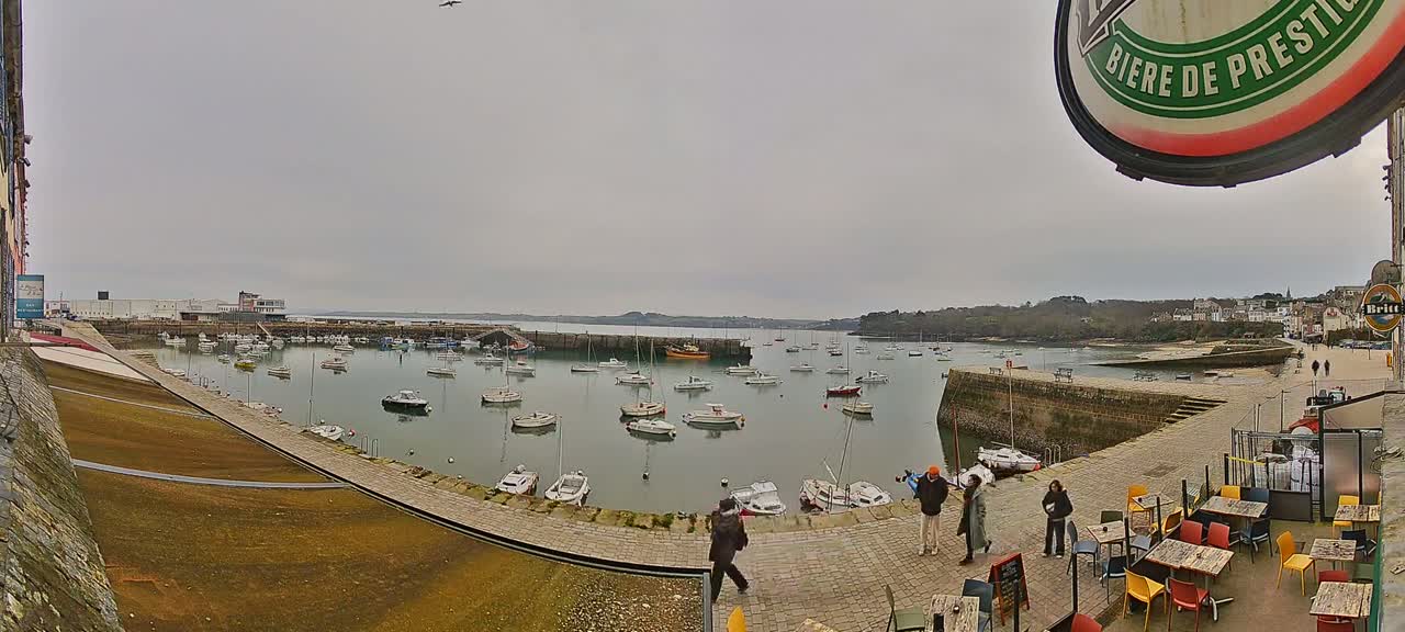 A harbor filled with numerous small boats is viewed from a slightly elevated position on an overcast day, with several people standing near an outdoor cafe in the foreground.