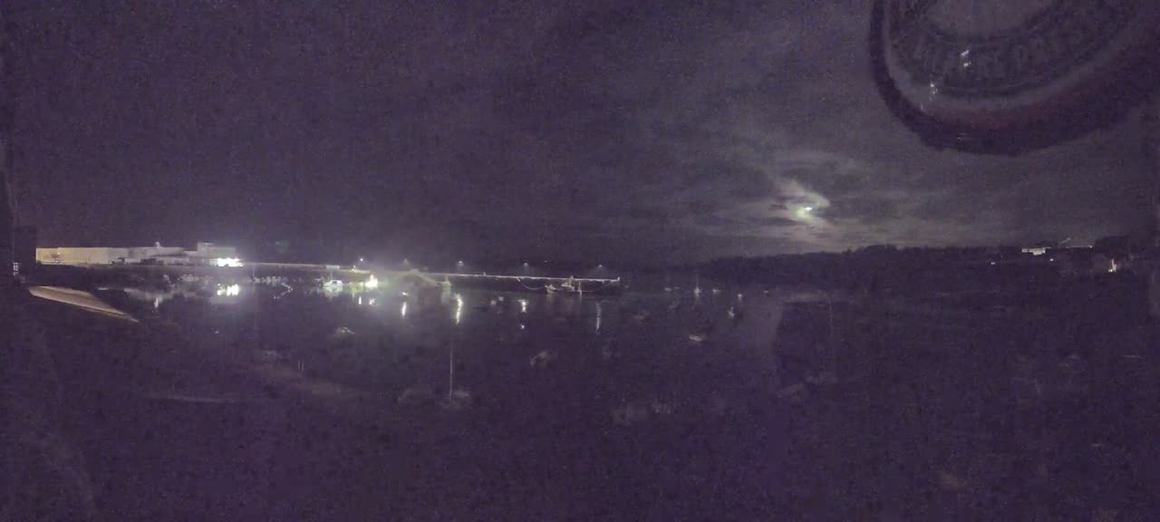 A nighttime view of a harbor with boats and lights, under a partly cloudy sky.