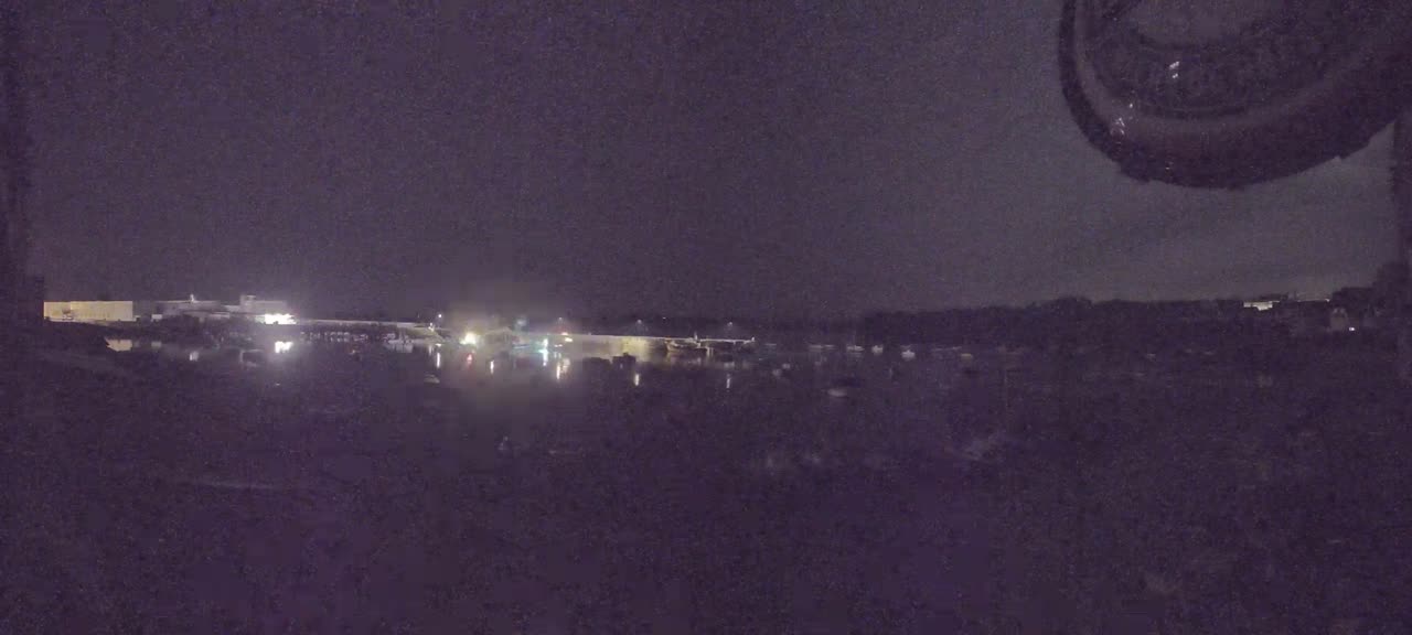 A nighttime view shows a body of water with dimly lit buildings and boats across the water under a dark sky.