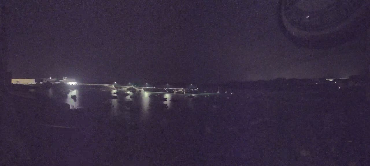 A nighttime view of a harbor with boats and lights reflecting on the water under a dark sky.