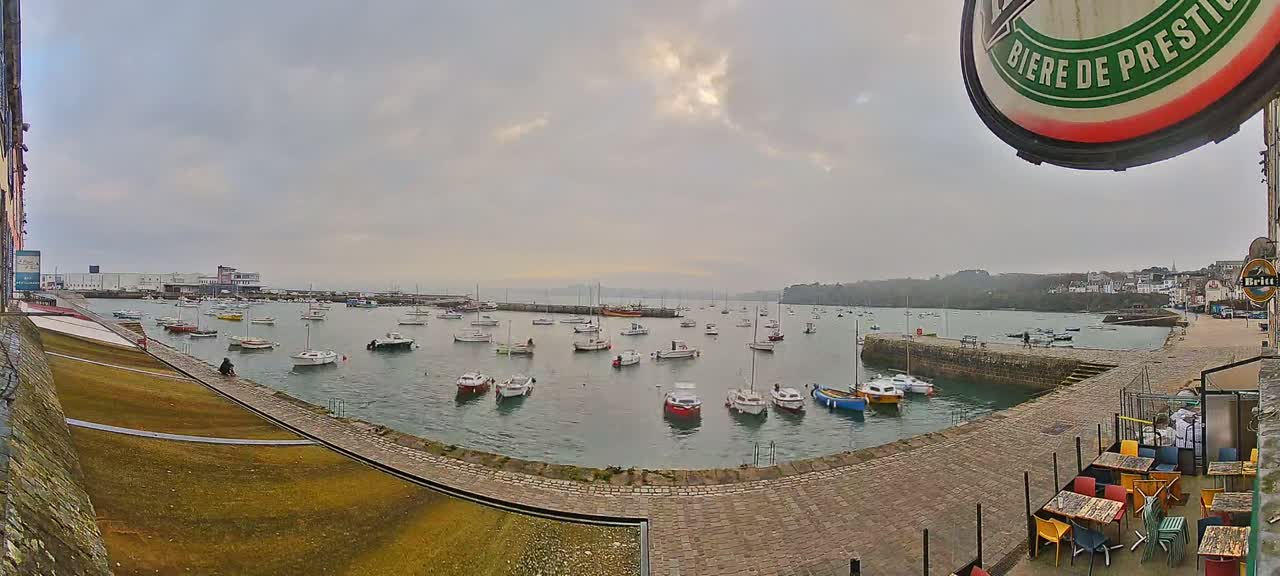 A harbor filled with numerous small boats is seen on an overcast day, with a paved walkway and outdoor seating area visible in the foreground.
