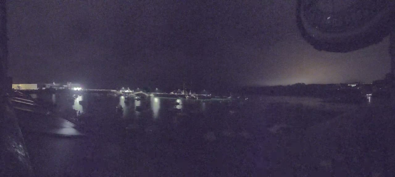 A nighttime view shows numerous boats moored in a harbor, illuminated by faint lights against a dark sky.