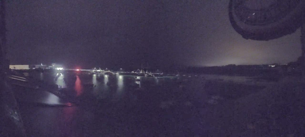 A nighttime view of a harbor filled with boats, illuminated by lights, under a dark, cloudy sky.