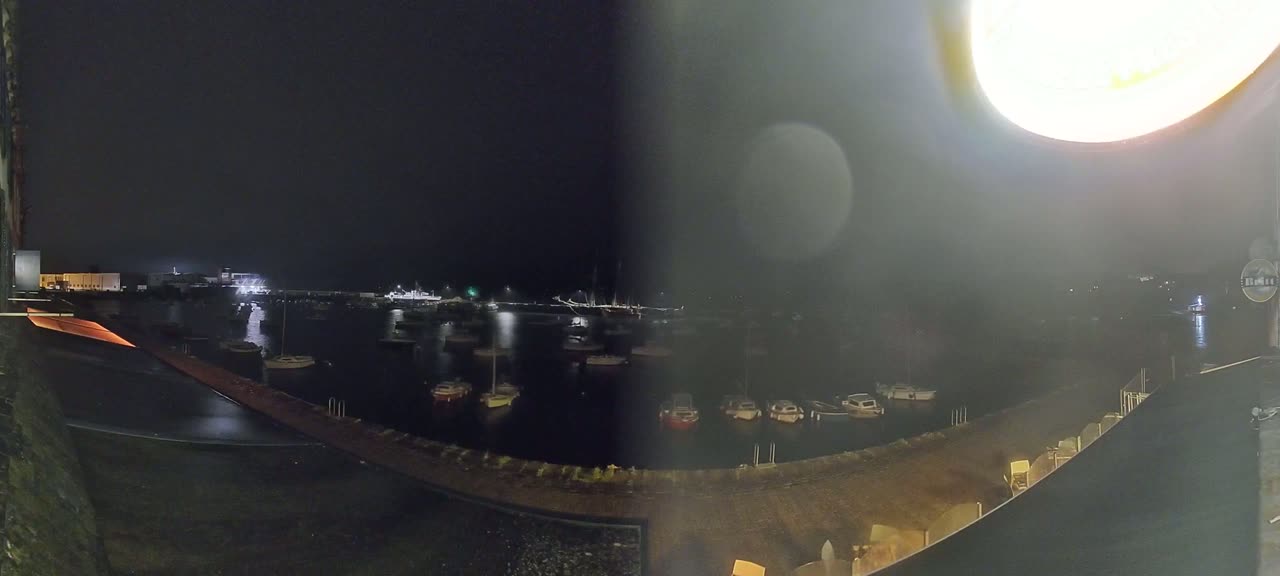A nighttime view of a harbor filled with boats, illuminated by lights from the shore and a bright light source in the upper right corner.