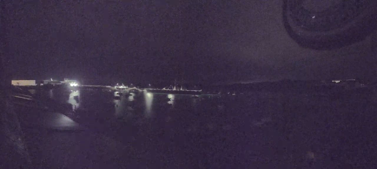 A nighttime view of a calm body of water with sparsely lit boats and buildings along the shore under a dark sky.