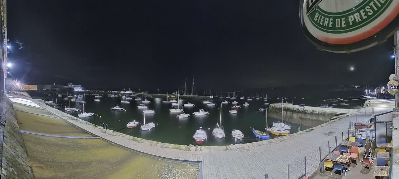 At night, numerous small boats are moored in a calm harbor, illuminated by artificial lights.