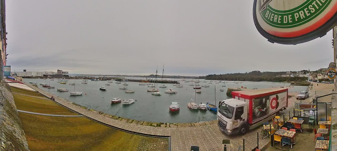 A harbor filled with numerous boats on an overcast day, with a delivery truck parked near an outdoor patio seating area.