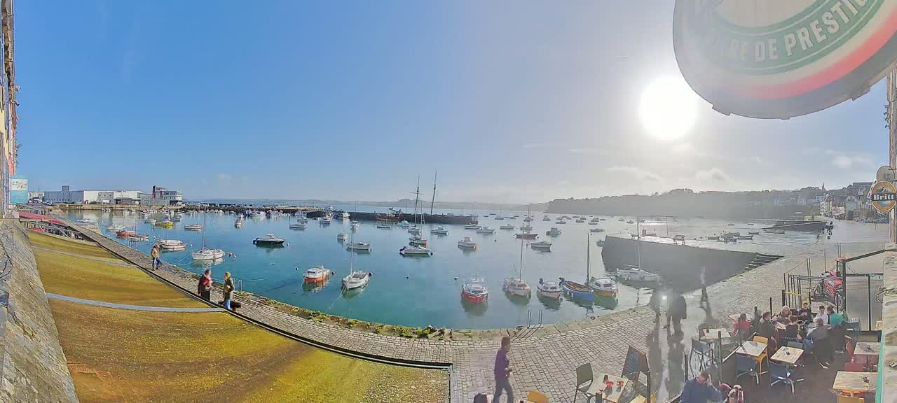 A sunny day overlooking a harbor filled with numerous sailboats and motorboats, with people walking along a stone walkway and sitting at outdoor tables near a building.