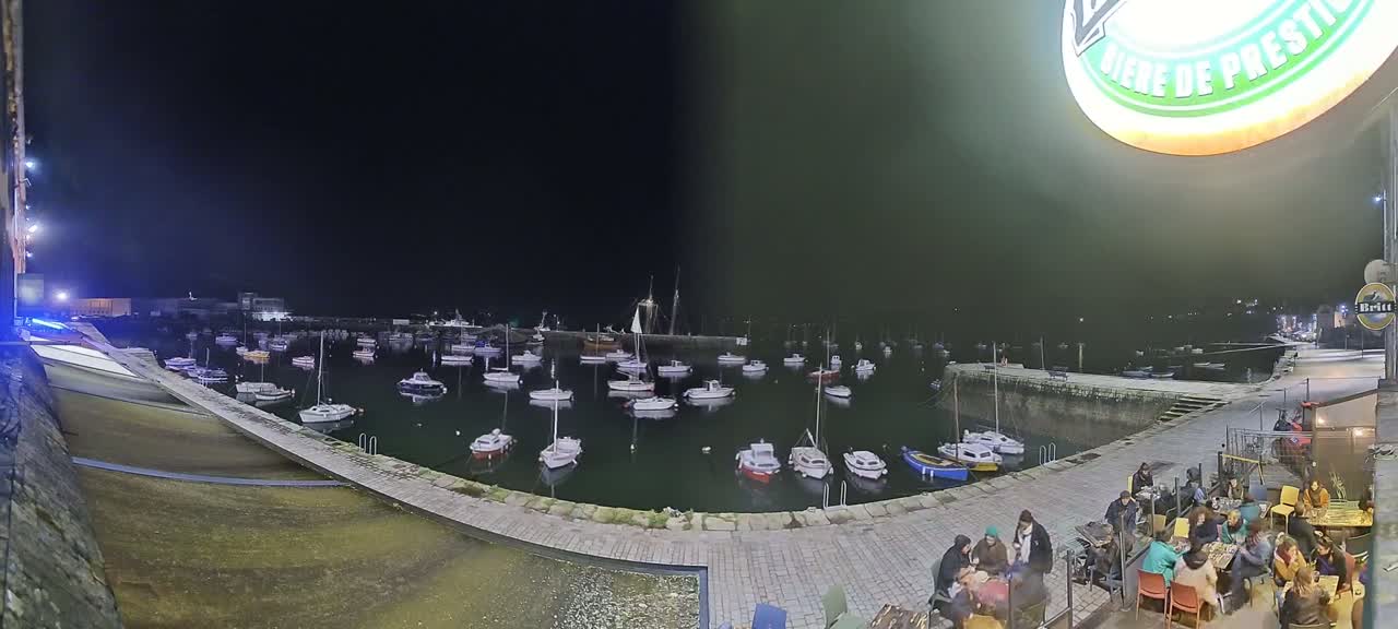 A nighttime harbor scene shows numerous small boats moored in calm water, with people sitting at outdoor tables near a stone waterfront.
