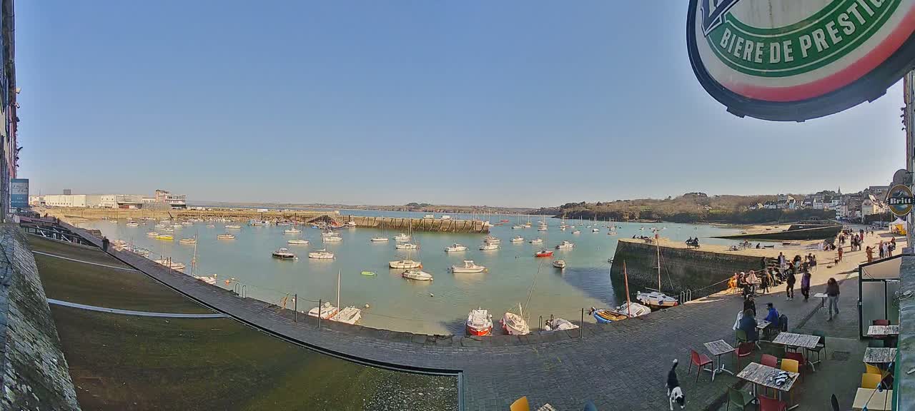 A sunny harbor scene shows numerous small boats moored in calm, shallow water, with a paved waterfront area featuring outdoor seating and people strolling along a seawall.