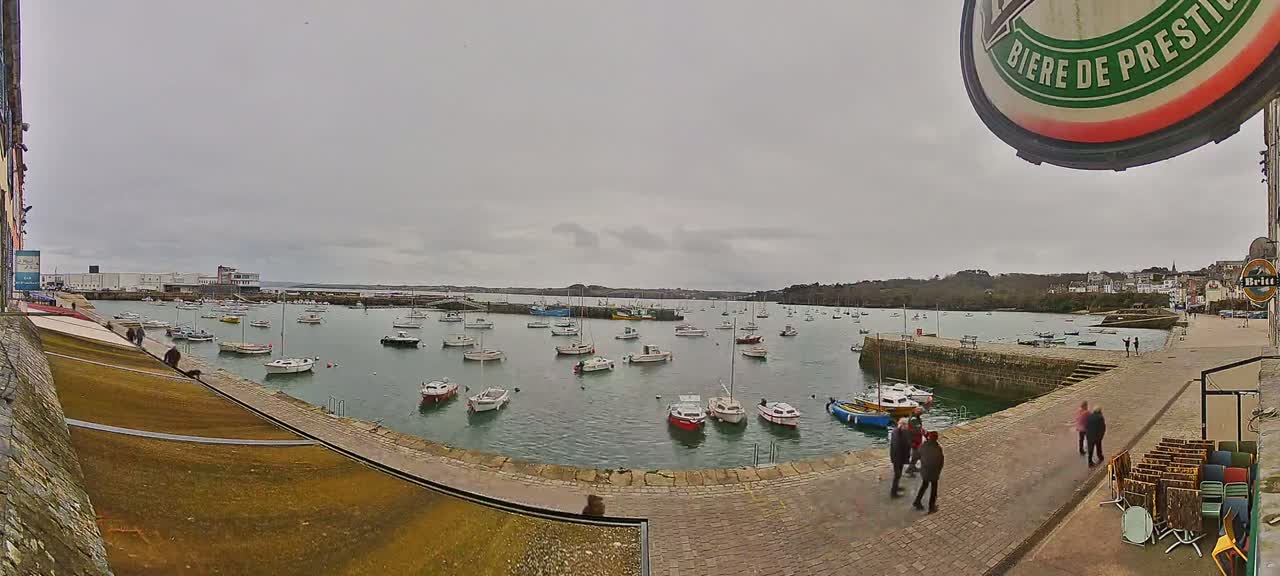 A harbor filled with numerous small boats is seen under an overcast sky, with several people walking along a paved area next to the water.