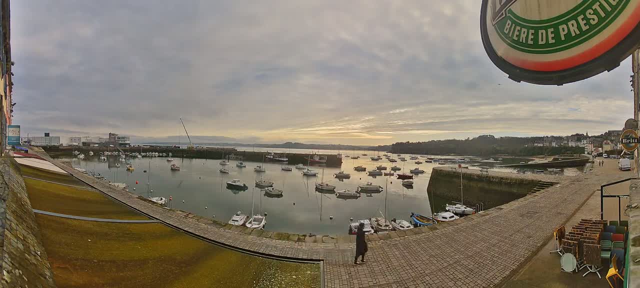 A calm harbor filled with numerous boats is viewed from a cobblestone walkway on an overcast day, with a sunset visible in the distance.