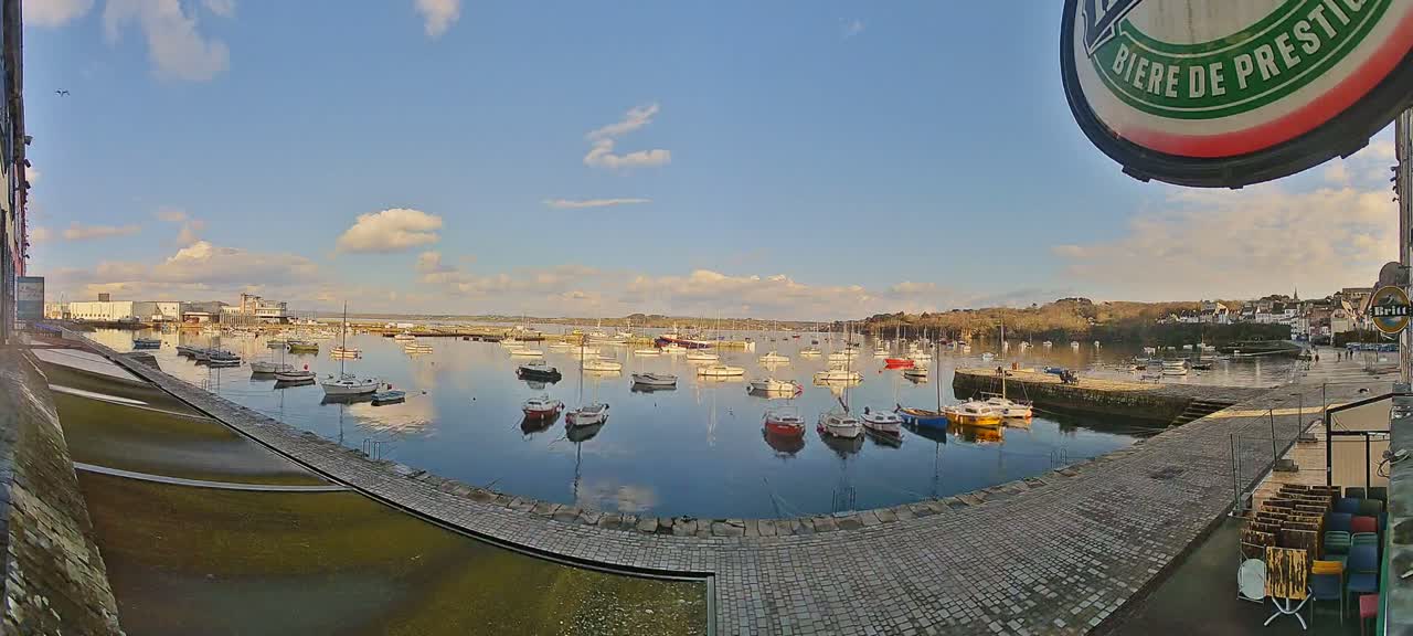 Numerous small boats are moored in a calm harbor under a partly cloudy, sunny sky.