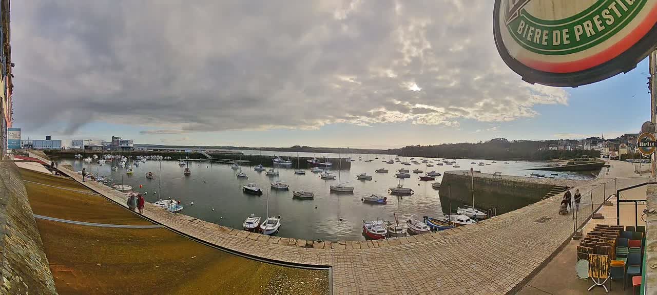 A harbor filled with numerous small boats is seen under a mostly cloudy sky, with a few people walking along a stone walkway.
