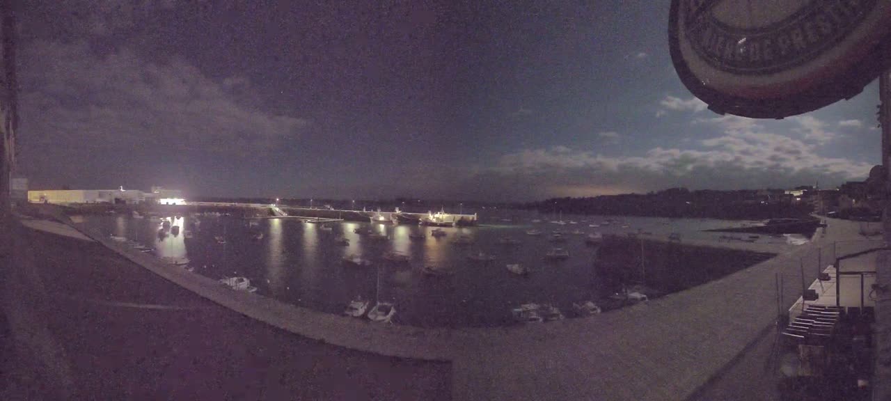 A nighttime harbor scene under a partly cloudy sky shows numerous boats moored in the calm water, illuminated by lights from the shore and a few boats.