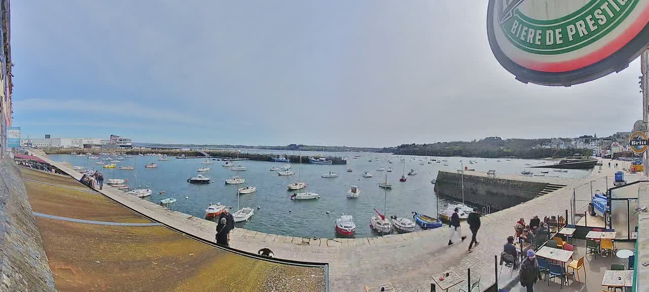 A harbor filled with numerous small boats on a partly cloudy day, with people walking along a stone quay and sitting at outdoor tables near buildings.