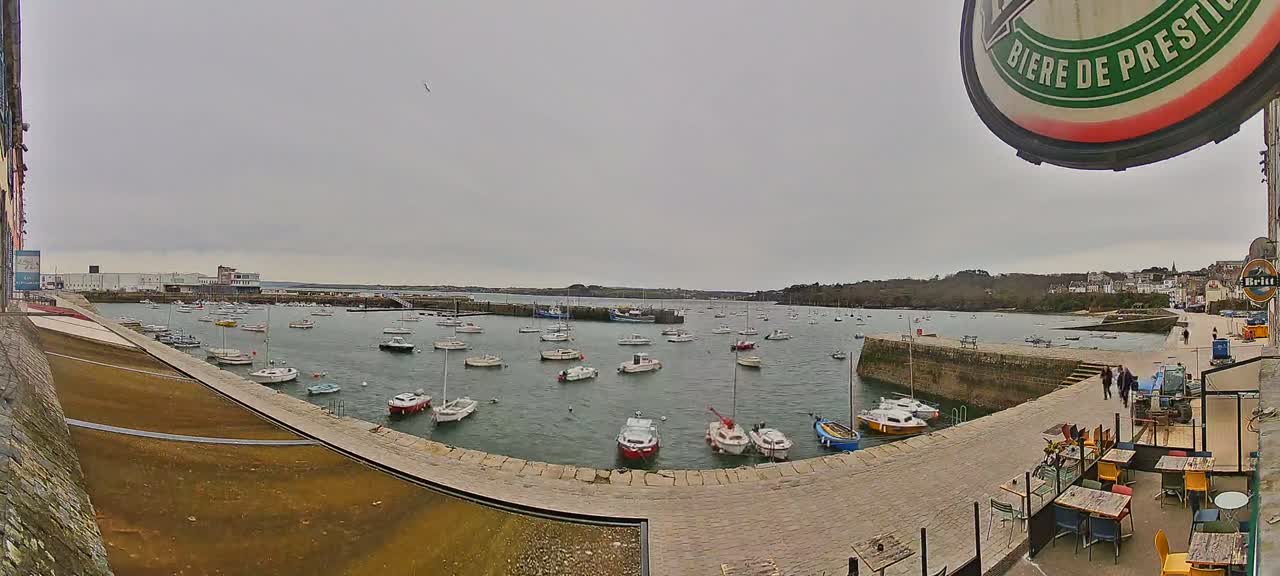 Many small boats are moored in a calm harbor on an overcast day, with a stone quayside and outdoor seating area visible in the foreground.